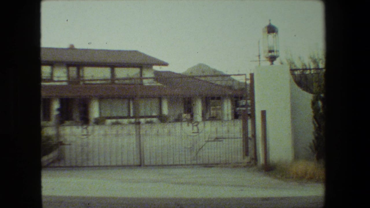 1981 Malibu home-movie frame showing a large two-story stucco residence behind a metal driveway gate with white pillars, lanterns, pavement, and shrubs.