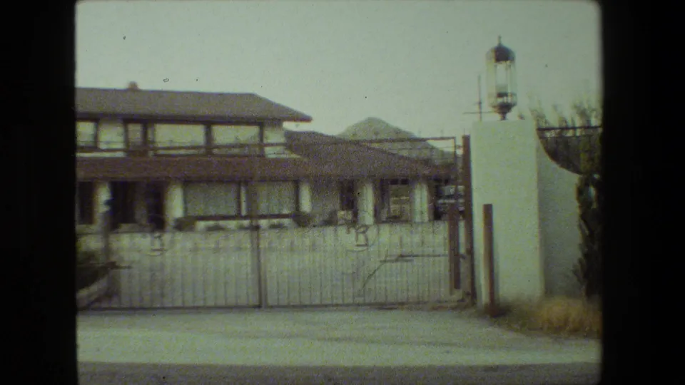 1981 Malibu home-movie frame showing a large two-story stucco residence behind a metal driveway gate with white pillars, lanterns, pavement, and shrubs.