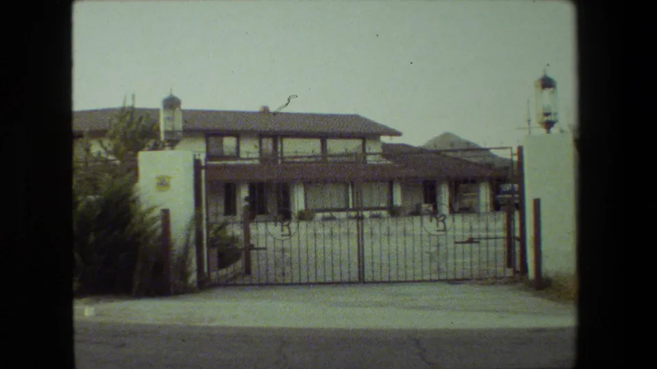 1981 Malibu exterior frame with a two-story stucco house, tiled roof, metal gate, white entrance pillars, lanterns, street pavement, and surrounding greenery.