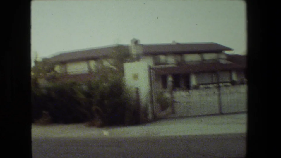 Soft 8mm frame from 1981 Malibu showing the gated stucco residence at a slight angle, with shrubs, pavement, a white pillar, and the tiled roof visible.