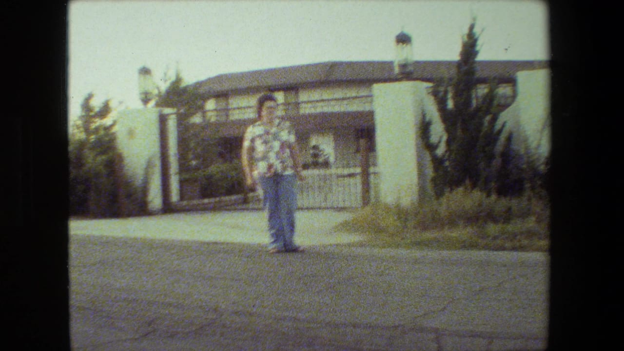 In the 1981 Malibu home-movie frame, a woman in a floral shirt and blue jeans stands on a road before a white stucco gate and tiled-roof house.