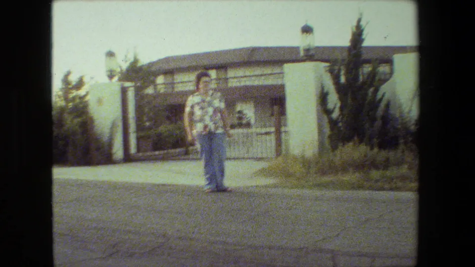 In the 1981 Malibu home-movie frame, a woman in a floral shirt and blue jeans stands on a road before a white stucco gate and tiled-roof house.
