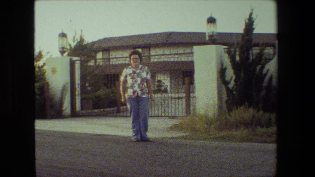 Woman standing farther right on the road with a stucco house and gate behind her.