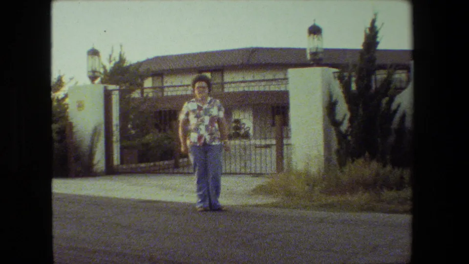 A woman wearing glasses, a patterned shirt, and jeans stands on a Malibu residential street with a metal gate, stucco wall, shrubs, and tiled roof behind her.