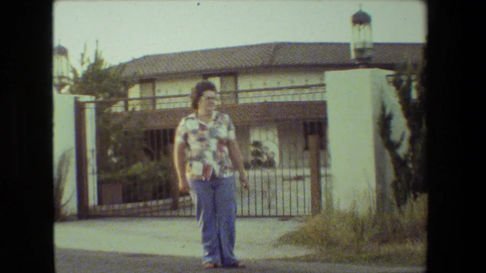 The woman appears closer in frame, standing on the street in front of the gated stucco residence with the tiled roof and landscaping visible behind her.