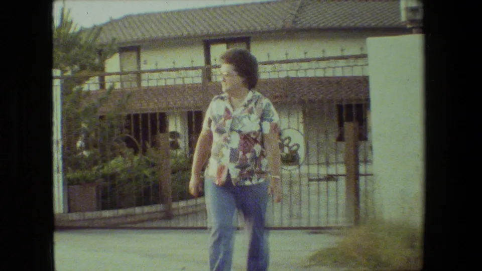 Near the end of the Malibu home-movie clip, the woman walks toward the camera while looking sideways, with the stucco columns, gate, barred windows, and garden plantings behind her.