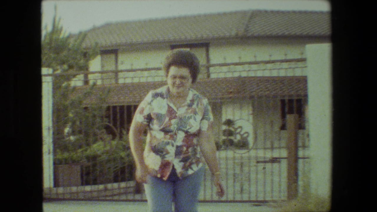 A woman in a floral short-sleeve shirt and blue jeans runs near a metal gate and white wall in 1981 Malibu home-movie footage.