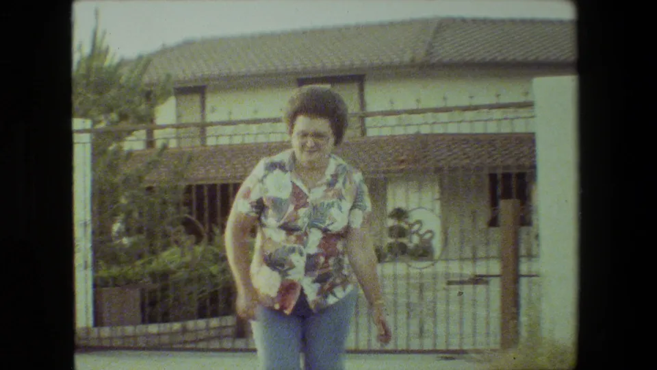 A woman in a floral short-sleeve shirt and blue jeans runs near a metal gate and white wall in 1981 Malibu home-movie footage.