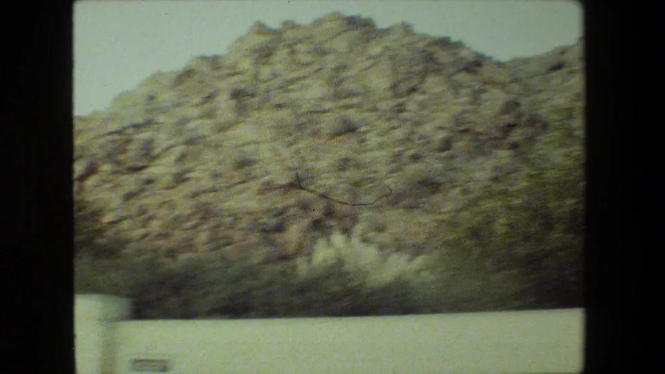 A softly focused handheld 8mm view shows a rocky hillside with sparse shrubs above a white wall in the Malibu residential setting.