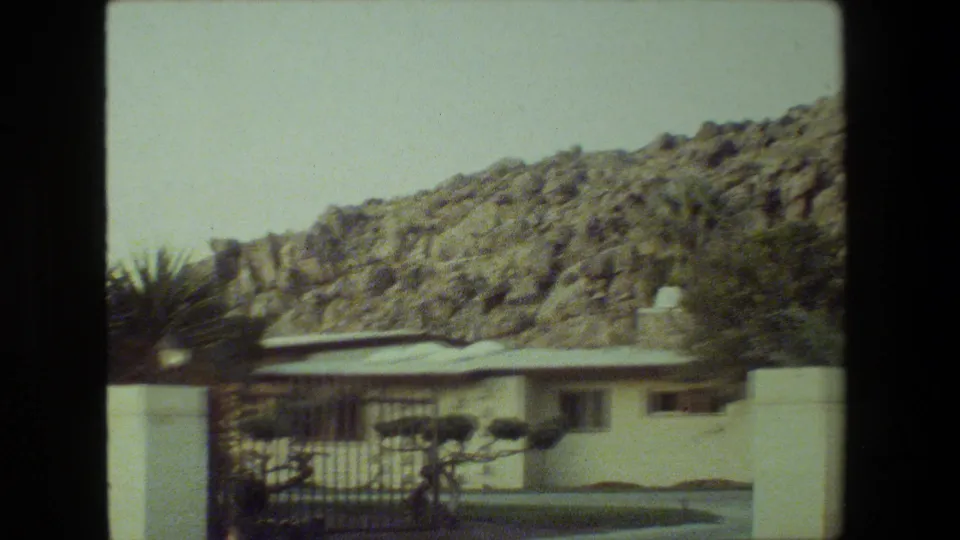 A low residential exterior sits below a rocky hillside, with a white wall, gate, shrubs, and sunlit yard visible in 1981 Malibu footage.