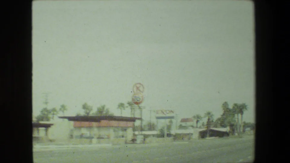 A soft, faded 8mm frame shows roadside businesses, palm trees, and a distant Exxon sign in Malibu, California, during the 1981 road-trip footage.