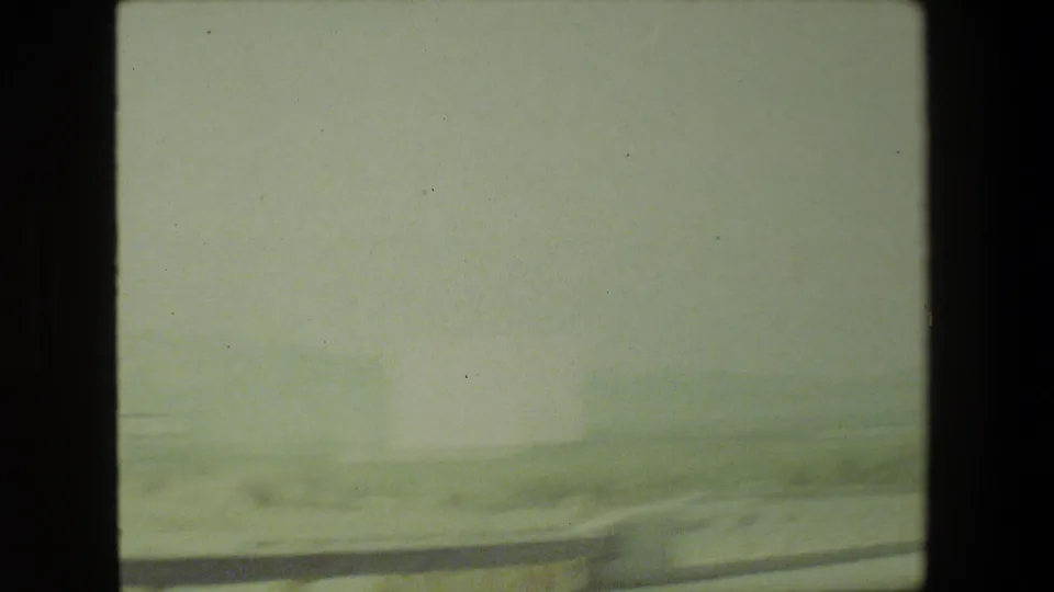 Bright, soft 8mm frame from the moving vehicle showing arid roadside ground and distant terrain through dark interior window framing.
