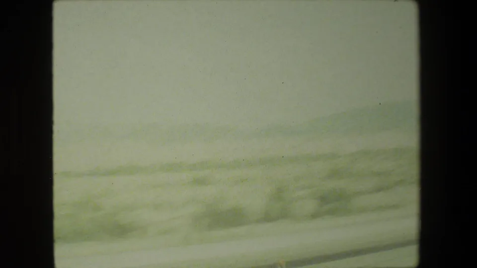 1981 Malibu home-movie frame showing motion-blurred roadside ground and low hills from a car-window perspective.