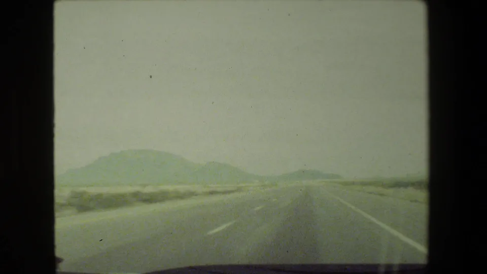 1981 8mm view from a moving vehicle on a hazy highway near Malibu, California, with distant mountains and lane markings visible.