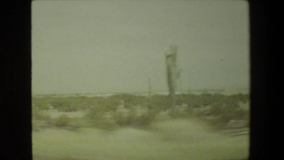 1981 8mm frame of a hazy arid roadside view with low scrub and distant cactus-like forms seen through motion blur.