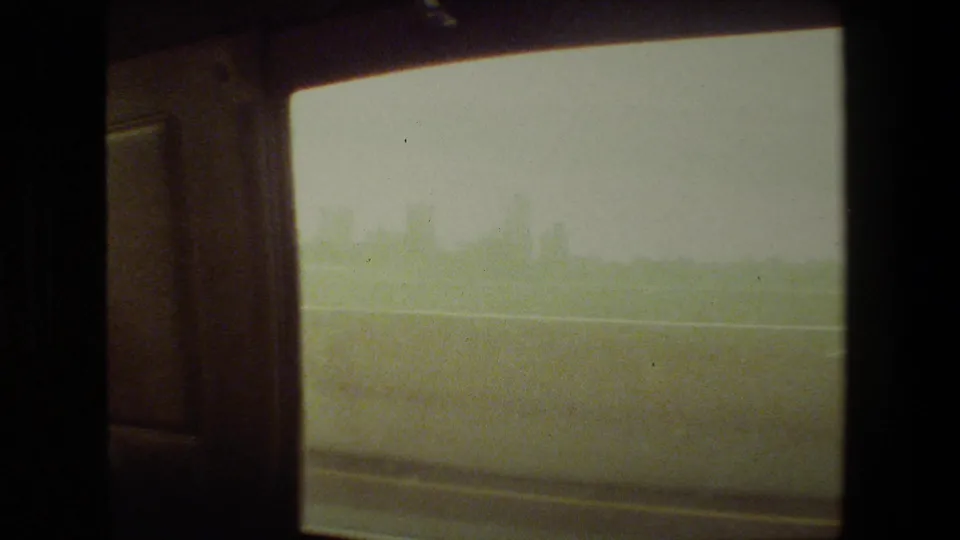 Soft 8mm frame from inside a moving vehicle, showing a dark interior edge, pale sky, roadside barrier, and blurred open landscape in the Malibu-area footage.