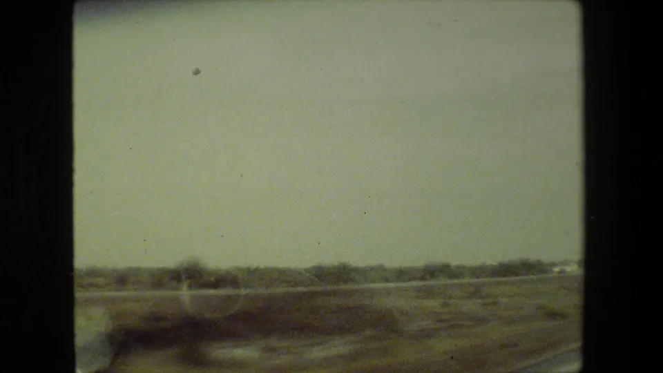 Final 1981 Malibu-area car-window view showing open arid ground, low vegetation, hazy sky, a dark lower-frame shape, and visible 8mm grain.
