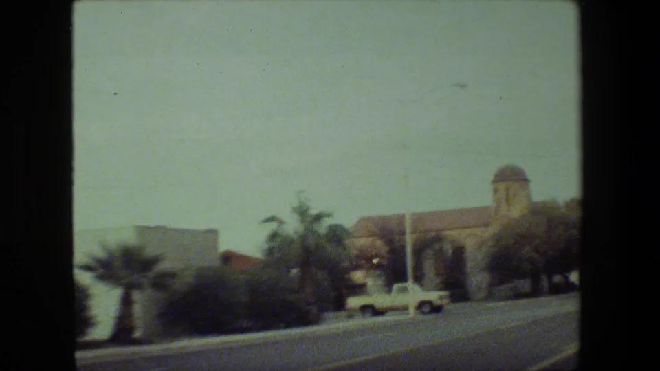 1981 Malibu-area street view with palm trees, parked or passing cars, and a low building with a dome, filmed from a moving vehicle on 8mm.