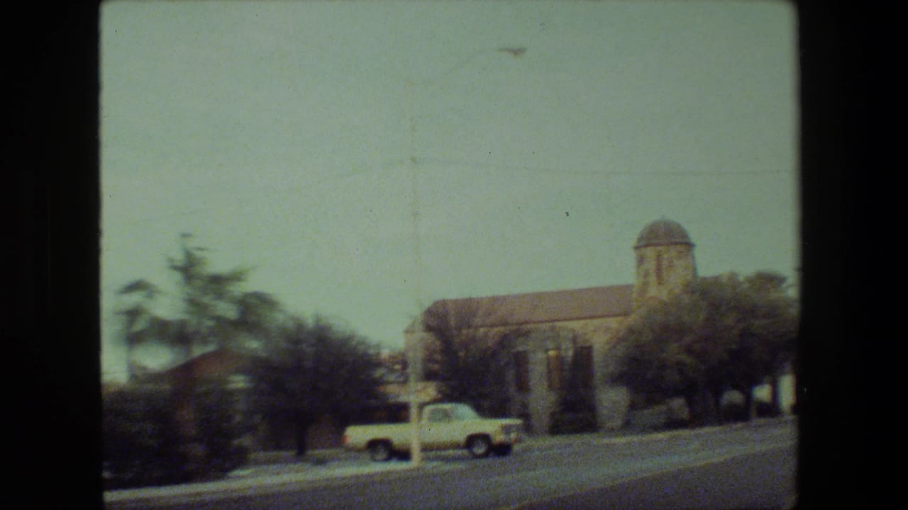 1981 Malibu 8mm frame from inside a moving vehicle, showing a stone church-like building with a small dome, palm trees, roadway, and a light-colored car passing in front.