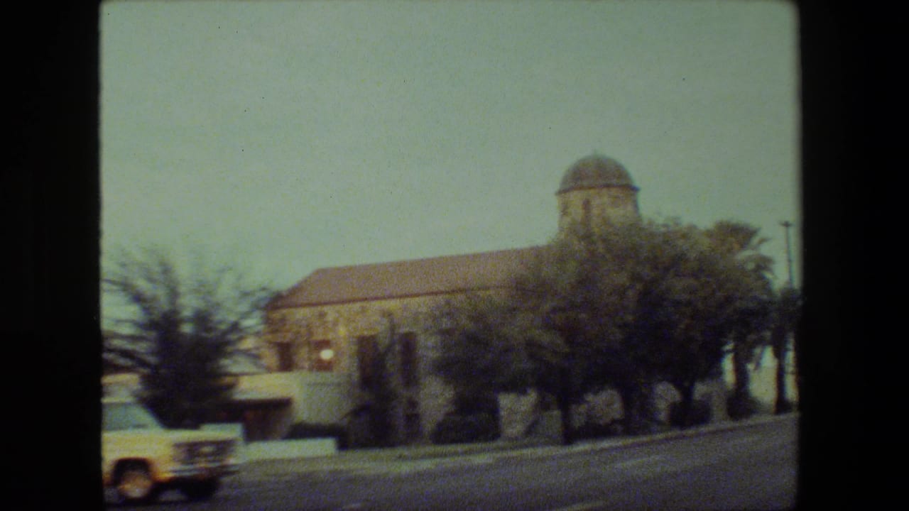 Yellow station wagon passes in front of a stone church-like building with palm trees along the road.