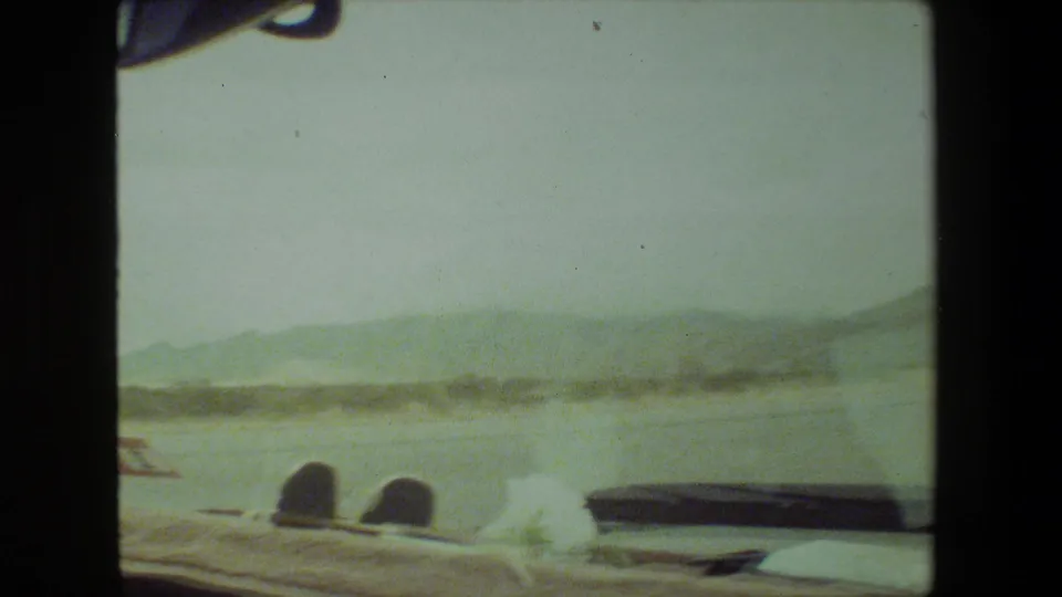 1981 Malibu passenger-view frame looking over the vehicle interior toward roadside structures, open terrain, distant hills, and a hazy sky.