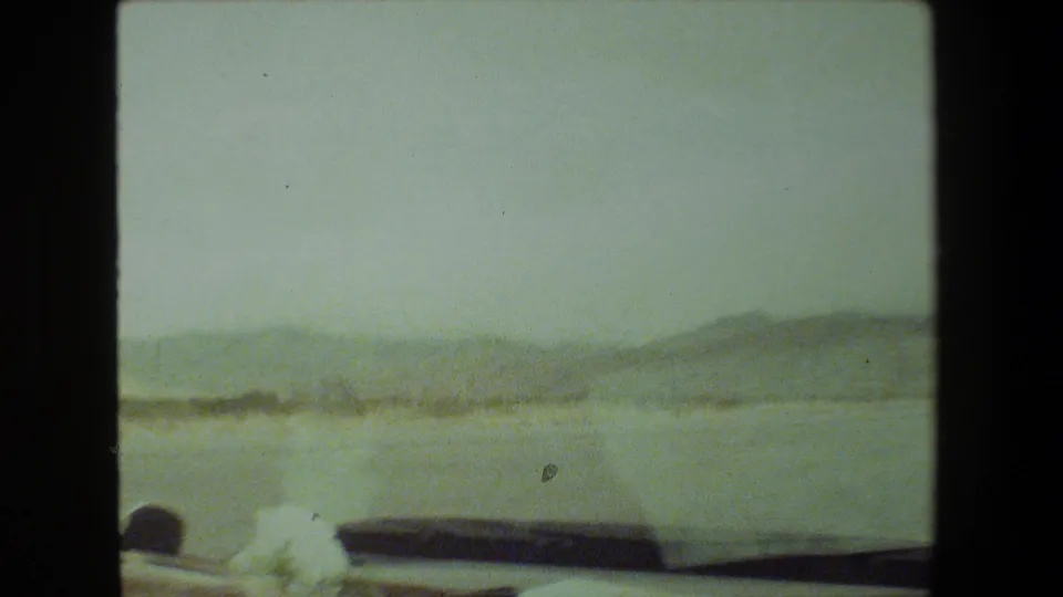1981 Malibu 8mm frame from a moving car, showing a dashboard edge, roadside low structures, pale open ground, distant hills, and a washed-out sky.