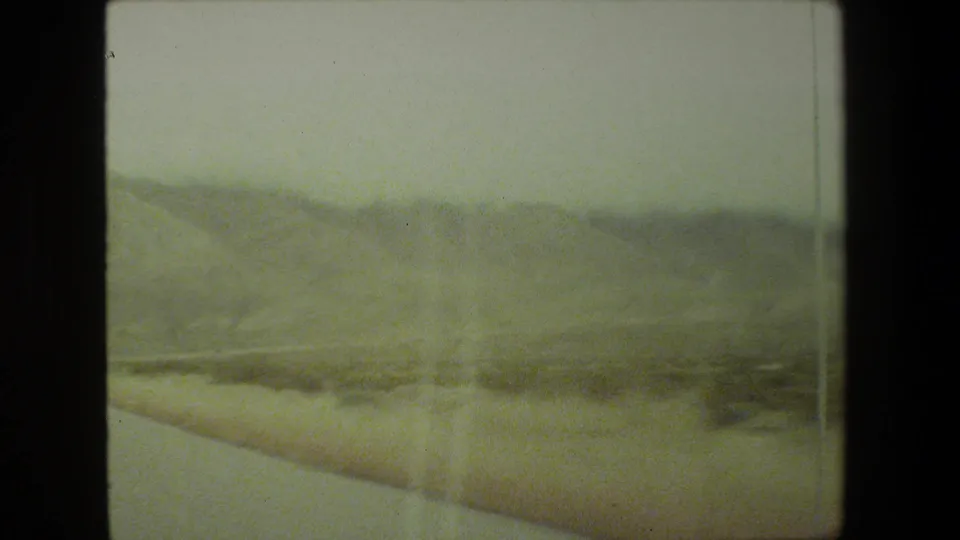 Passenger-view 8mm frame from the Malibu road sequence, showing roadside brush, a pale road edge, and distant hazy hills under an overcast sky.