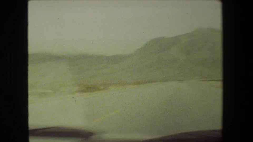 Final frame of the Malibu highway clip, with hazy hills, a pale road or shoulder, and dark blurred vehicle foreground visible in the lower edge.