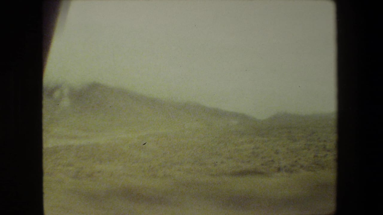 1981 Malibu home-movie frame showing dry rolling hills and roadside vegetation through a softly blurred moving-vehicle view.