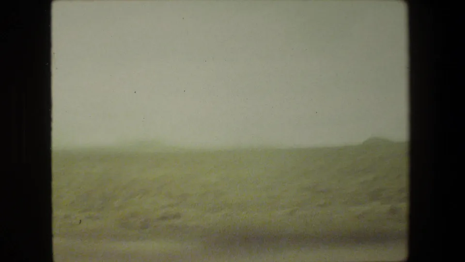 Moving-vehicle frame from 1981 Malibu showing blurred roadside scrub, low hills, and a bright hazy sky on 8mm film.