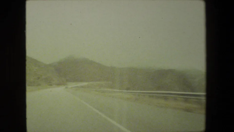 1981 Malibu road-trip frame showing a curving highway, metal guardrail, road shoulder, and dry hills ahead through a moving vehicle window.