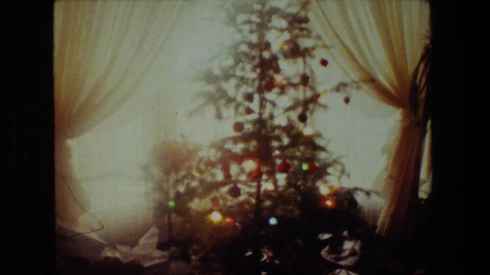 The home-movie view shifts to a decorated Christmas tree in a private Malibu interior, with glowing lights, ornaments, sheer curtains, and wrapped gifts below.