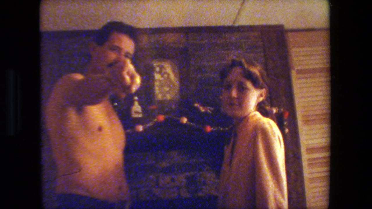 In a 1981 Malibu home interior, an adult man and woman stand near a decorated brick fireplace in warm holiday light.