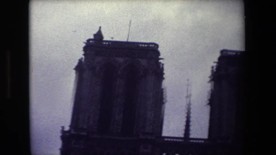 1981 Paris 8mm frame with a close handheld view of Notre-Dame's left tower, central spire, and the edge of the opposite tower against gray sky.