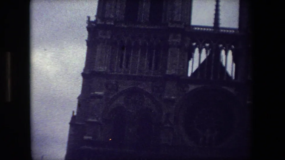 1981 Paris 8mm frame showing more of Notre-Dame Cathedral's facade, including twin towers, central spire, and dark Gothic arch forms beneath a cloudy sky.