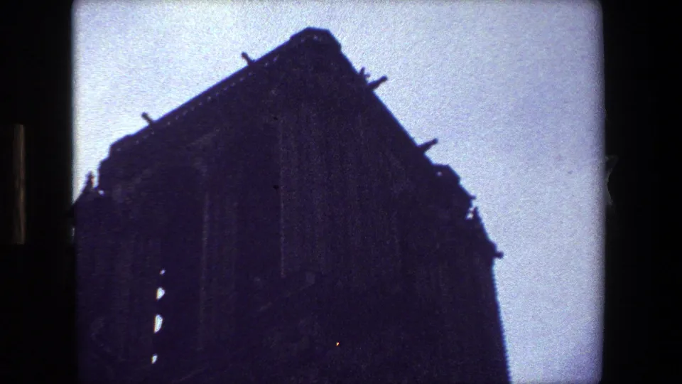 1981 Paris frame showing a tall shadowed architectural facade with roof projections and a pale sky, rendered with soft 8mm grain and muted color.