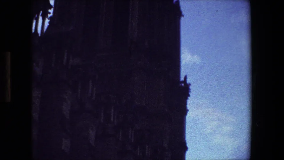 1981 Paris 8mm frame with the cathedral facade filling most of the view, its stonework mostly in shadow beside pale blue sky and cloud.