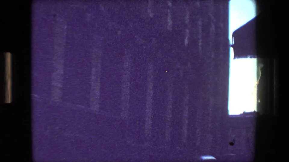 Purple-tinted home-movie frame showing a dark interior edge, a broad wall or facade surface, and a bright vertical window opening in the Paris street-view sequence.