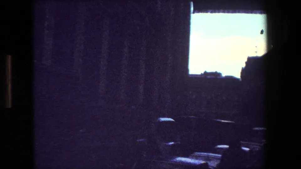 From the supplied 1981 Paris 8mm clip, a dim interior looks out through a bright window toward a shadowed street area with indistinct cars or urban forms below.