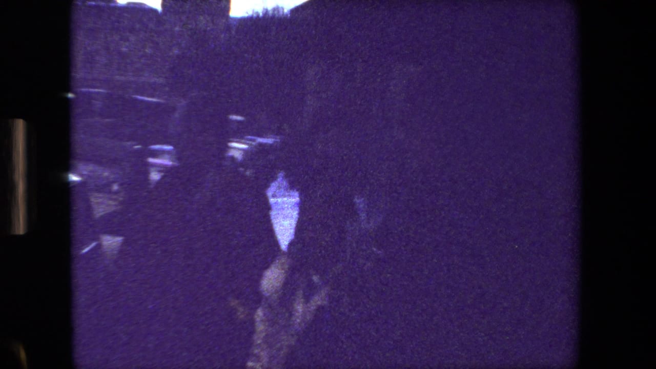 Dark silhouetted figure near parked cars and building shadows in a purple-tinted street view.