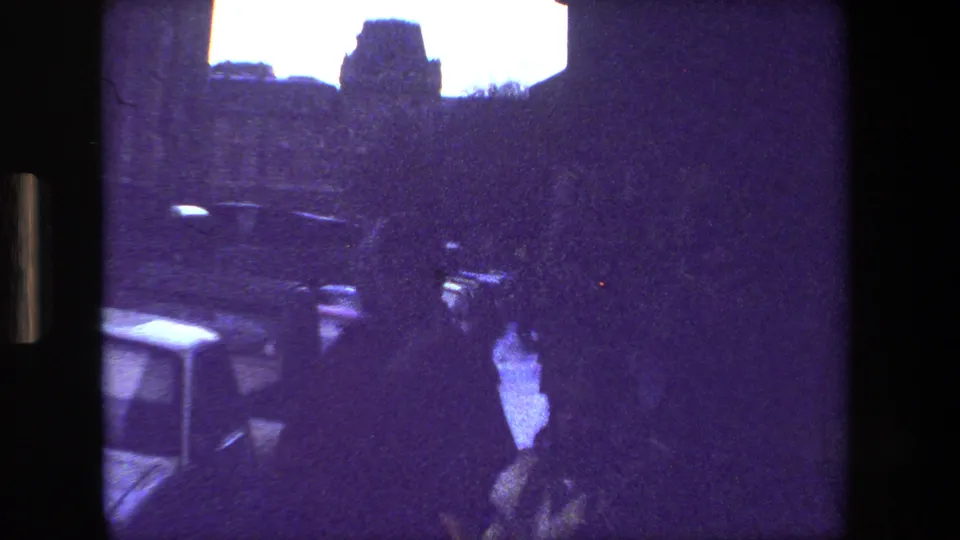 A grainy 8mm frame from Paris in 1981 appears to show a person in silhouette near parked cars and a narrow street or courtyard, with strong purple color shift.