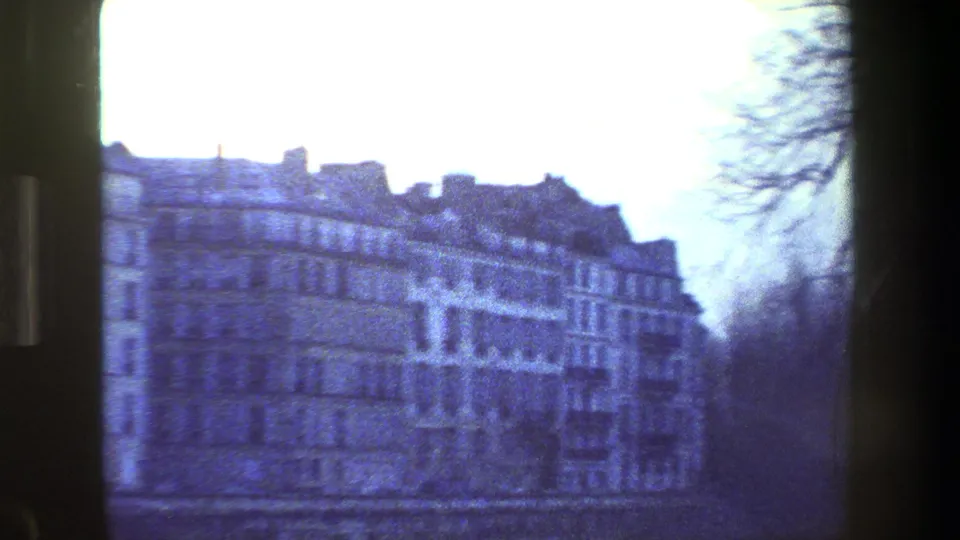 1981 Paris home-movie view across a river toward pale multi-story buildings, framed by a railing or fence and winter trees.