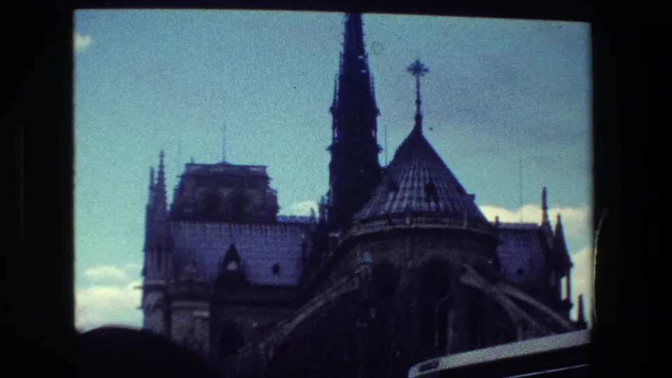 Notre-Dame Cathedral in Paris appears in a grainy 1981 8mm frame, with Gothic roof forms, a tall spire, cloudy sky, and dark fence framing.