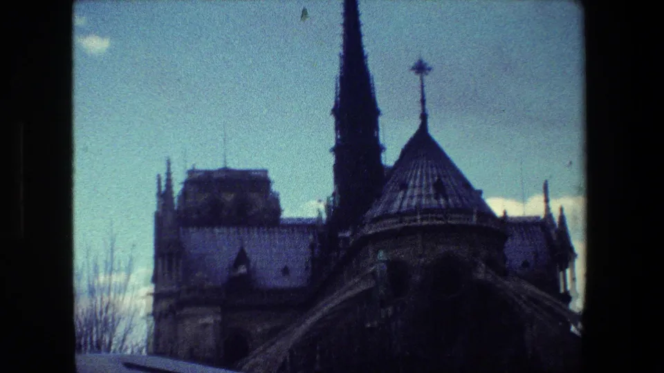 A cool-toned 1981 Paris 8mm frame shows Notre-Dame Cathedral's spire and roofline beyond a dark vertical fence frame, with sky and bare branches nearby.