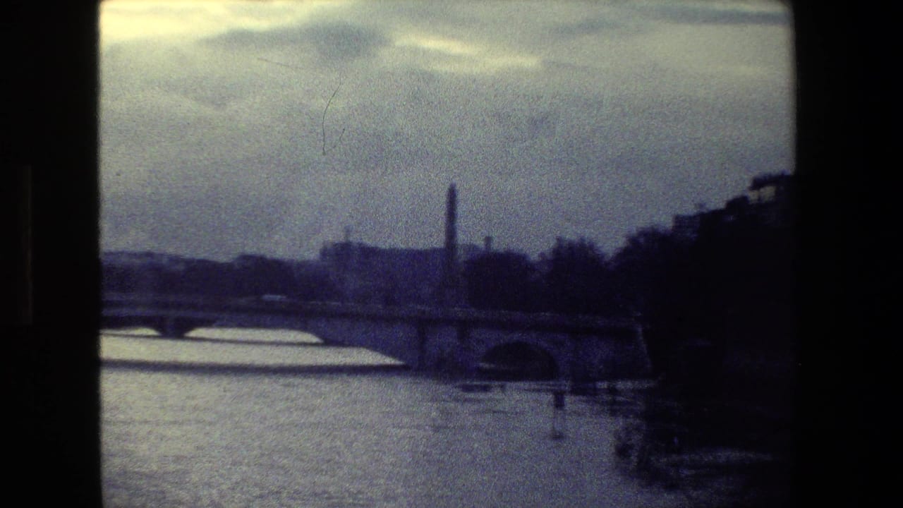 1981 Paris frame looking over the Seine toward a stone bridge, riverside embankment, and distant vertical monument-like form under a muted sky.