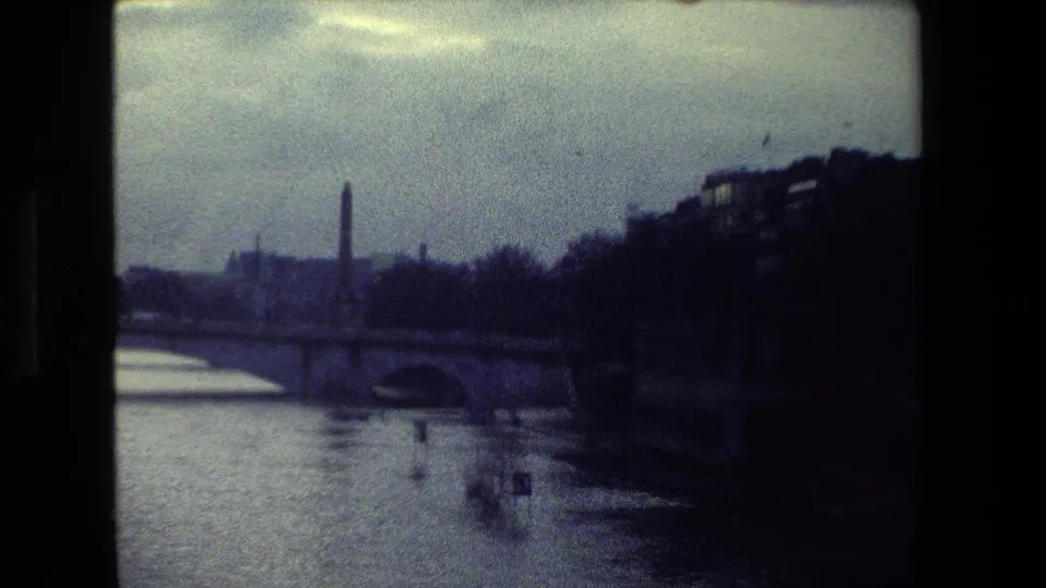 1981 Paris home-movie view across the Seine, with bridge arches, a shadowed right bank, bare trees, and a pale overcast sky.
