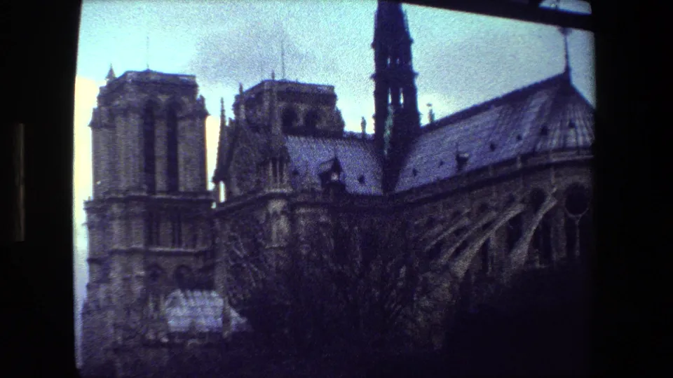 Vintage 8mm frame from Paris showing a Gothic cathedral exterior with twin towers, a central spire, roof details, and dark tree silhouettes under a cloudy sky.