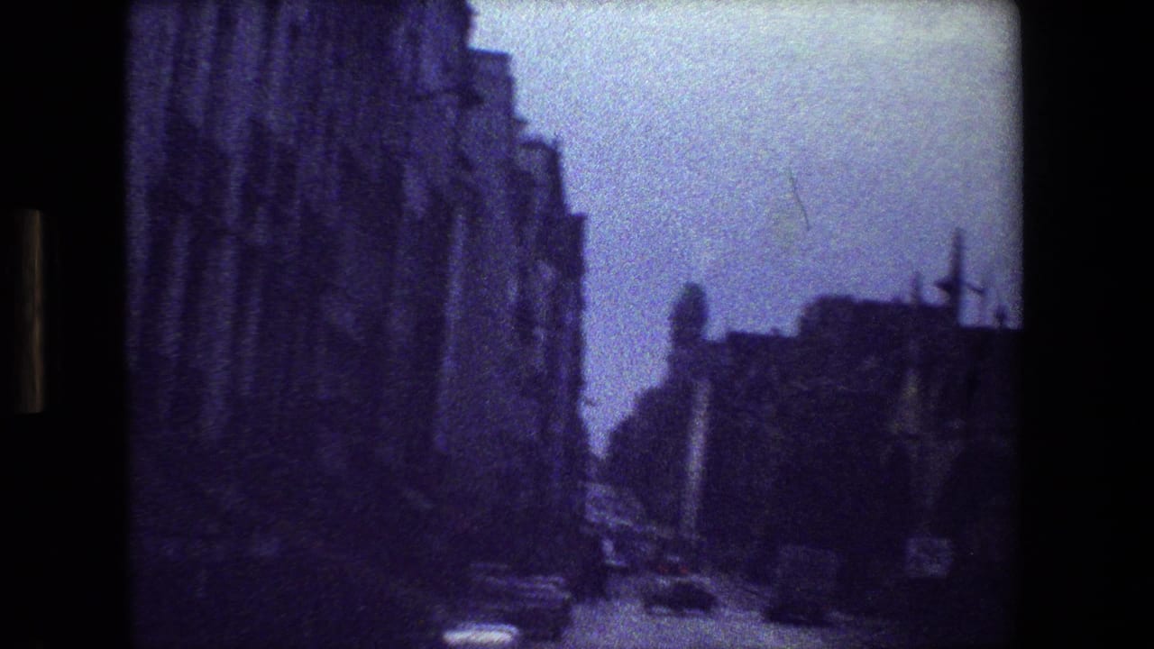 Dim street view beside tall stone buildings with cars on the road.