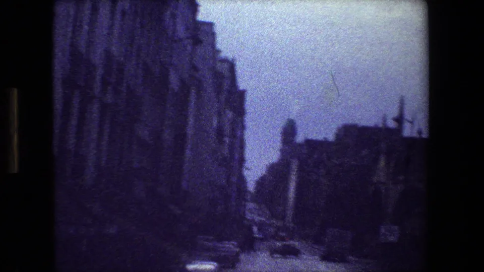 Grainy 8mm Paris street view with tall stone buildings, a narrow roadway, small cars, and a dark foreground edge from the film frame.