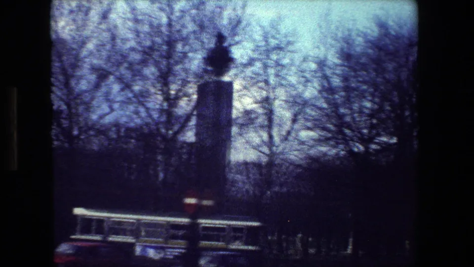 Paris street view from 1981 8mm footage with bare trees, a passing bus, cars, and a tall street fixture softened by motion blur and film grain.